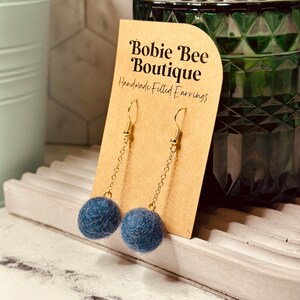 Op de afbeelding: Handgemaakte vilten oorbellen met een gouden ketting en haak. De oorbellen hebben een donkerblauwe vilten bal. Ze worden gepresenteerd op een bruine kaart met de tekst "Bobie Bee Boutique Handmade Felted Earrings."