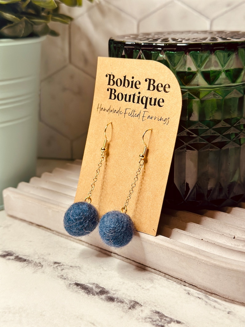 Op de afbeelding: Handgemaakte vilten oorbellen met een gouden ketting en haak. De oorbellen hebben een donkerblauwe vilten bal. Ze worden gepresenteerd op een bruine kaart met de tekst "Bobie Bee Boutique Handmade Felted Earrings."