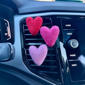 May include: Three felt heart-shaped car air fresheners in a car's air vent. The hearts are red, pink, and light purple.