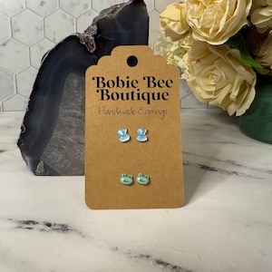 May include: A pair of handmade earrings featuring a blue bunny and a green frog design. The earrings are displayed on a brown card with the text "Bobbie Bee Boutique Handmade Earrings".
