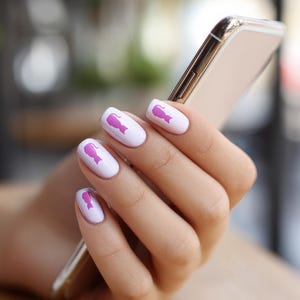 May include: A hand holding a gold smartphone, showcasing a manicure with white nails. Each nail features a pink design. The background is blurred, suggesting an outdoor setting, with a hint of greenery and a table.