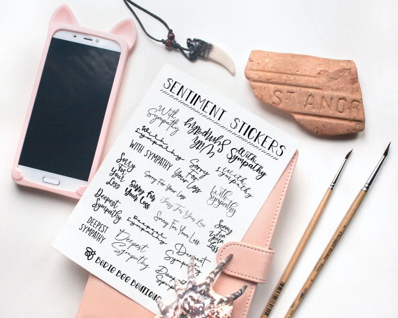 Puede incluir: Una hoja de papel blanco con pegatinas de caligraf&iacute;a negra que dicen "With Sympathy", "Sorry for Your Loss" y "Deepest Sympathy". El papel est&aacute; sobre un diario encuadernado en cuero rosa con una concha de mar en la parte superior.