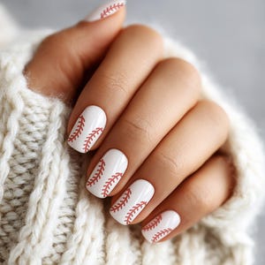 May include: Close-up of a hand with white nail polish, each nail decorated with a baseball stitch design in red. The hand is resting on a cream-colored knitted sweater.