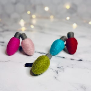 May include: A collection of colorful felt Christmas light ornaments in pink, light pink, teal, red, and green. Each light has a gray base. The ornaments are arranged on a white surface with bokeh lights in the background.