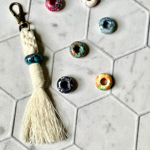 May include: A white tassel keychain with a silver key ring and a blue, green, and black patterned bead. The keychain is surrounded by six colorful ceramic beads with various patterns.