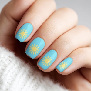 May include: A hand with light blue nail polish, each nail adorned with a yellow sun design. The hand is resting on a white knitted sweater, with a neutral background.