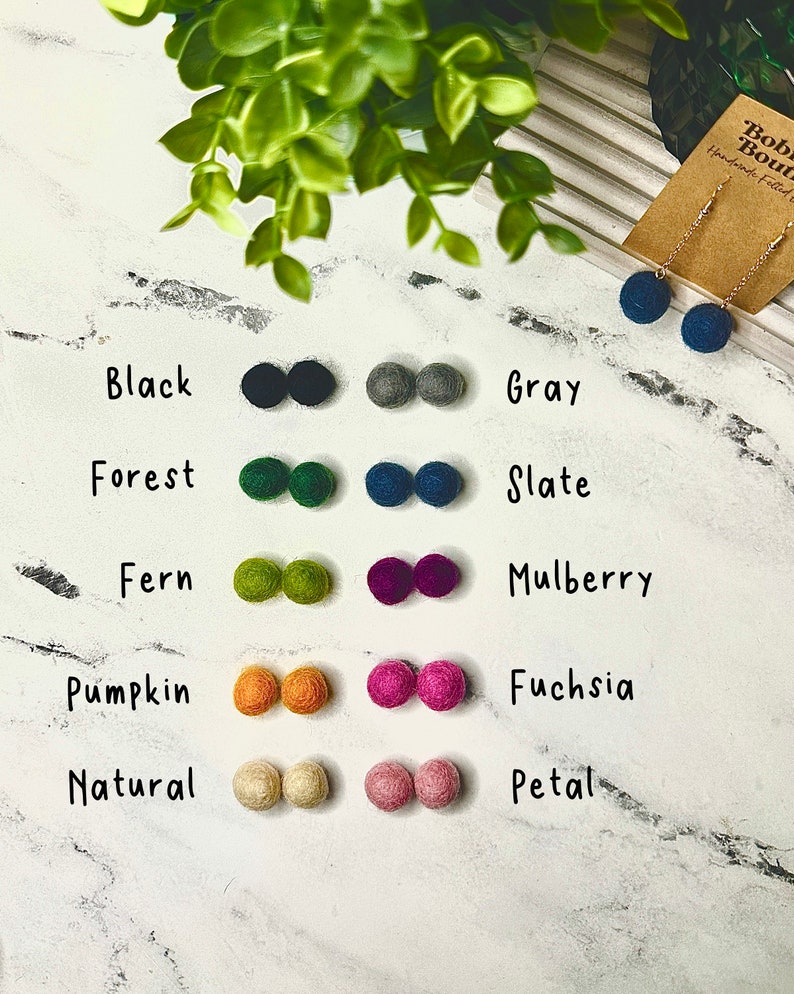 Op de afbeelding: Viltbal oorbellen en viltballen in diverse kleuren, waaronder zwart, grijs, bosgroen, leisteenblauw, varengroen, moerbei, pompoen, fuchsia, naturel en bloembladroze. Er is ook een paar leisteenblauwe oorbellen te zien.