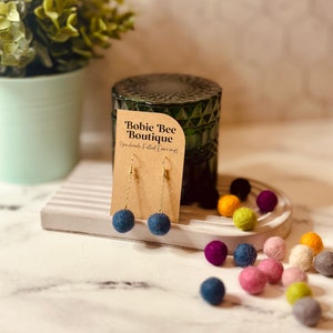 Op de afbeelding: Handgemaakte vilten oorbellen met marineblauwe bolvormige hangers aan gouden kettingen. De oorbellen worden getoond op een kaartje met de tekst "Bobie Bee Boutique". Kleurrijke vilten ballen in diverse tinten liggen rond de oorbellen.