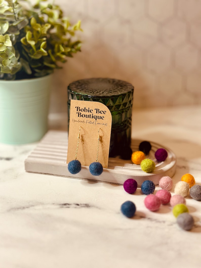 Op de afbeelding: Handgemaakte vilten oorbellen met marineblauwe bolvormige hangers aan gouden kettingen. De oorbellen worden getoond op een kaartje met de tekst "Bobie Bee Boutique". Kleurrijke vilten ballen in diverse tinten liggen rond de oorbellen.