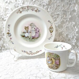 May include: A white ceramic plate and mug set featuring illustrations from Beatrix Potter's "Tom Kitten." The plate has a central image of a cat and a figure in a pink dress. The mug depicts rabbits. Floral accents and text adorn both pieces.