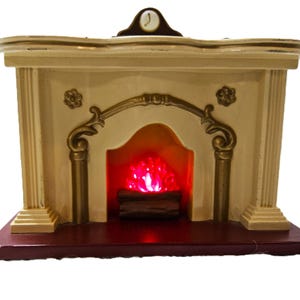 May include: A cream-colored decorative fireplace mantel with gold-toned accents, featuring a central archway, two side columns, and a small clock face on top. A glowing red light simulates a fire over brown logs on a dark red base.