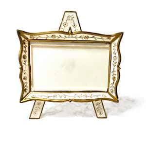 May include: A decorative white and gold picture frame on a small easel. The frame has a rectangular opening with a white backing. The frame is embellished with gold trim and floral designs.