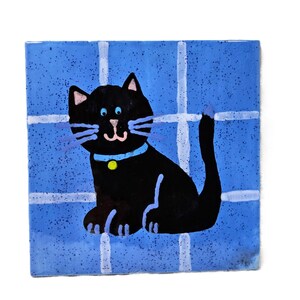 May include: A square ceramic tile featuring a hand-painted black cat with blue eyes, a light blue collar with a yellow tag, and long blue whiskers. The cat is set against a speckled blue background divided by a white grid pattern.