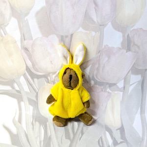 May include: A small brown teddy bear wearing a bright yellow bunny costume with long white-lined ears. The bear is posed against a backdrop of white and light gray tulip flowers.