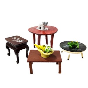 May include: A collection of four miniature tables including a dark brown rectangular table with white inlay, a round reddish-brown table with a small gray elephant figurine, a rectangular brown table with a yellow bowl of fruit, and a round black table.