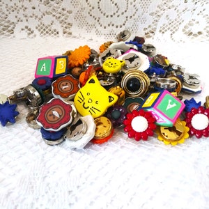 May include: A collection of colorful plastic buttons with various designs, including letters, animals, and flowers. The buttons are arranged on a white lace background.