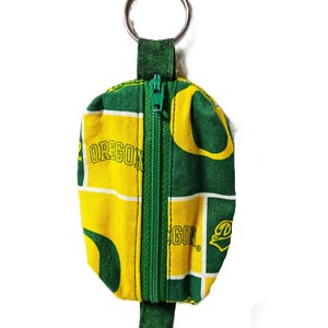May include: A small, zippered pouch with a key ring, featuring a green and yellow pattern with the word "OREGON" printed on it. The pouch has a green zipper and a green fabric loop for the key ring.