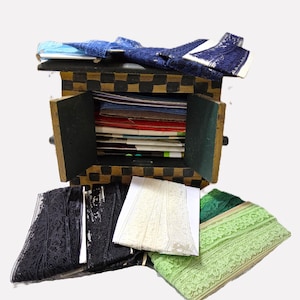 May include: A wooden chest with a black and gold checkered pattern, open to reveal stacks of paper and lace. Various colors of lace, including black, white, and green, are displayed around the chest. Blue lace is on top of the chest.