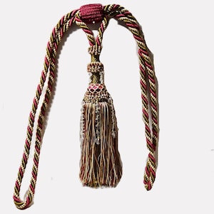 May include: An ornate curtain tieback with a tassel and braided cord. The tassel features a burgundy, gold, and brown color scheme, accented with clear beads. The braided cord is a mix of burgundy, gold, and cream.