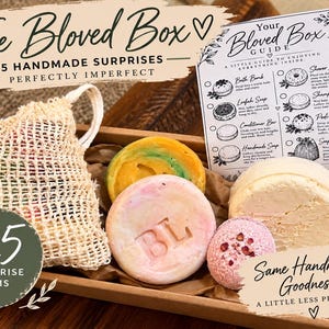 Peut inclure: Une boîte en carton rectangulaire contenant cinq articles de bain faits main, dont un sac à savon en maille beige, un savon rond jaune et vert, un savon rond rose avec BL en relief, une petite bombe de bain rose et un grand article rond blanc.