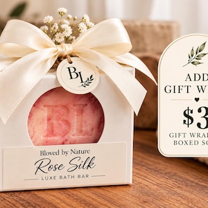 Peut inclure: Une boîte cadeau carrée blanche avec une découpe circulaire révélant un savon rose gaufré avec BL. La boîte présente un nœud en ruban de satin crème, une petite étiquette ronde avec BL et une étiquette indiquant Bloved by Nature Rose Silk Luxe Bath Bar.