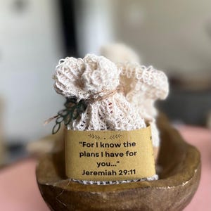May include: A small, off-white, textured fabric pouch tied with twine, featuring a brown paper band printed with the text, "For I know the plans I have for you..." followed by the scripture reference "Jeremiah 29:11" in black lettering.