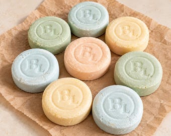 May include: Eight round solid shampoo bars in various pastel colors including light blue, sage green, peach, and pale yellow. Each bar features a circular embossed design with the letters BL in the center, arranged in a cluster.