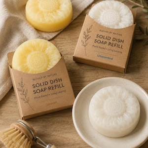 May include: Two round yellow and white solid dish soap refills in brown cardboard boxes labeled BLOVED BY NATURE SOLID DISH SOAP REFILL Plant-Based Long Lasting Lemon and Unscented, with two additional soap bars and a wooden dish brush.