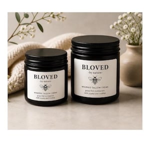 Peut inclure: Deux pots noirs de crème de suif fouettée "BLOVED", un hydratant nourri à l'herbe avec du jojoba et de l'églantier. Les pots ont des couvercles noirs et des étiquettes blanches avec une illustration d'abeille. Le fond comprend des fleurs et un tissu de ton neutre.