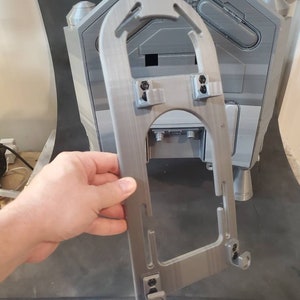 Könnte beinhalten: Ein grauer 3D-gedruckter Kunststoffrahmen mit einer rechteckigen Öffnung. Der Rahmen hat eine gebogene Oberseite und zwei kleine Laschen an der Unterseite.