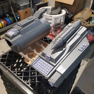 May include: Two 3D printed grey plastic props, one resembling a futuristic weapon with a handle and a barrel, and the other a smaller device with a keypad and a nozzle.