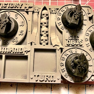 May include: A grey plastic game board with the words "Victory", "Command", "Turn", and "Sec Objs" printed on it. There are four dials with fleur-de-lis symbols and a row of skulls.