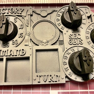 May include: A gray plastic game board with a skull and wings design. The board has multiple dials with numbers and symbols. The board has the words "VICTORY", "COMMAND", "SEC OBJS", and "TURN" printed on it.