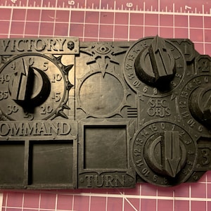 以下が含まれることがあります： 黒いゲームボードで、ダイヤルとテキストがあります。ボードには「Victory」、「Command」、「Turn」と書かれたダイヤルがあります。追加のダイヤルには「Sec Objs」とラベルが付けられています。ボードには、頭蓋骨と目のデザインがあります。