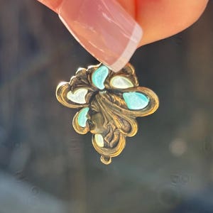 May include: A vintage-style pendant with a floral design. The pendant is made of gold-toned metal and features a profile of a woman's face. It is adorned with turquoise and white accents. The pendant is held up by a person's hand.
