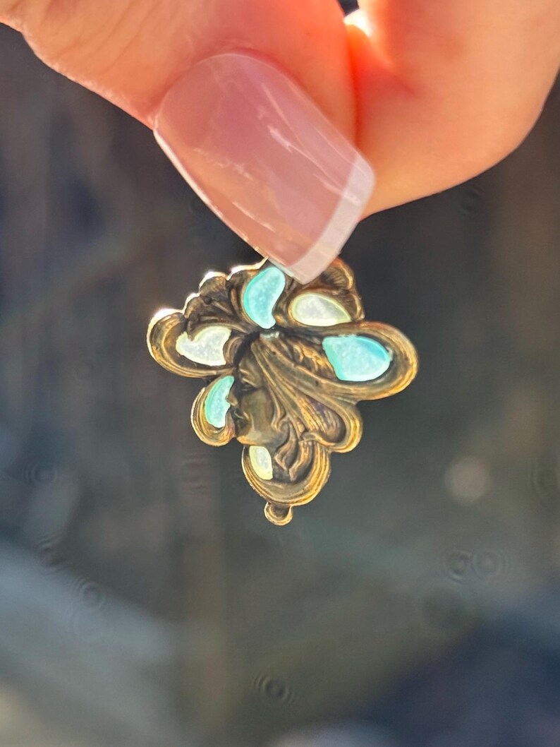 May include: A vintage-style pendant with a floral design. The pendant is made of gold-toned metal and features a profile of a woman's face. It is adorned with turquoise and white accents. The pendant is held up by a person's hand.