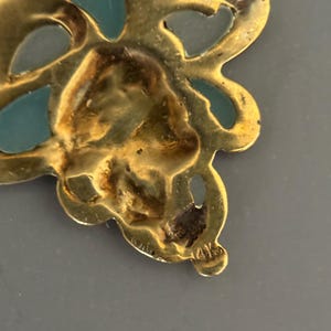 May include: Close-up of a gold-toned pendant with a floral design. The pendant features teal-colored enamel accents and is marked with "14K". The intricate details and color contrast create a vintage aesthetic.