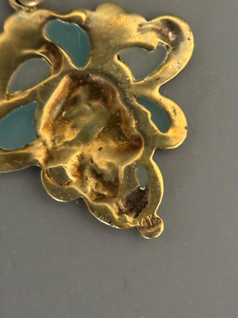 May include: Close-up of a gold-toned pendant with a floral design. The pendant features teal-colored enamel accents and is marked with "14K". The intricate details and color contrast create a vintage aesthetic.
