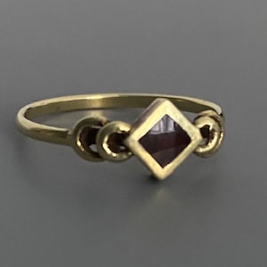 May include: A gold ring with a dark red square gemstone set in the center. The band of the ring has a decorative design.