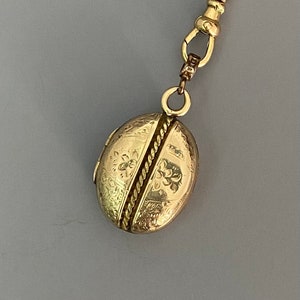 May include: A gold locket necklace with an oval-shaped locket featuring an intricate floral design. The locket is closed and has a rope-like design down the center.