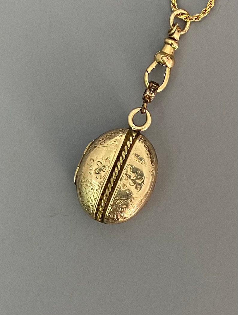 May include: A gold locket necklace with an oval-shaped locket featuring an intricate floral design. The locket is closed and has a rope-like design down the center.