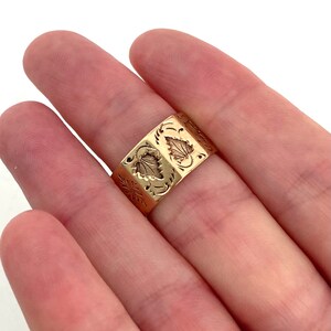 May include: A gold band ring with an intricate floral design. The ring is worn on a person's finger.