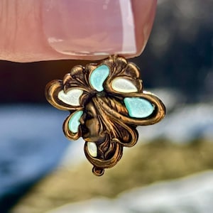 May include: A vintage bronze-toned pendant featuring a woman's profile. The pendant has a floral design with light blue and white enamel accents. The piece is held up by a person's finger.