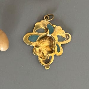 May include: A gold-colored pendant with a floral design and turquoise accents. The pendant has a loop at the top for a chain. The pendant is on a gray surface.