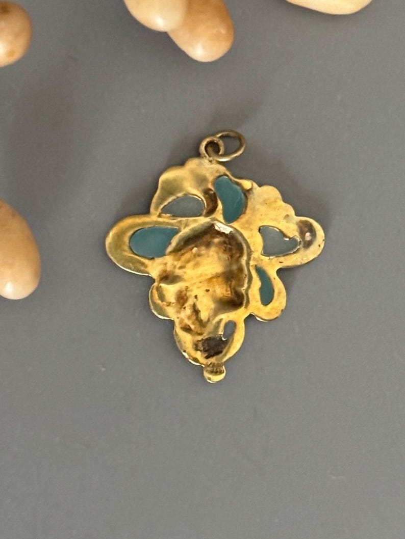 May include: A gold-colored pendant with a floral design and turquoise accents. The pendant has a loop at the top for a chain. The pendant is on a gray surface.