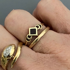 May include: Three gold rings on a finger. The top ring has a black square gemstone. The middle ring is a simple band. The bottom ring is a snake design with a small diamond.