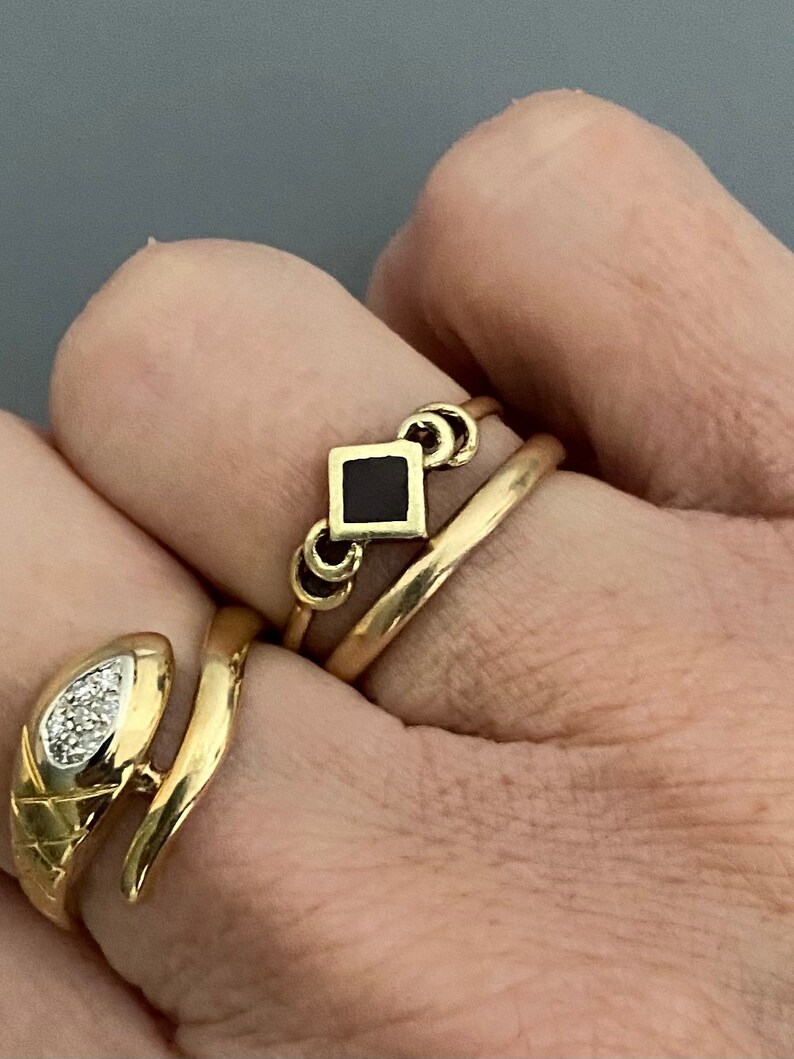 May include: Three gold rings on a finger. The top ring has a black square gemstone. The middle ring is a simple band. The bottom ring is a snake design with a small diamond.