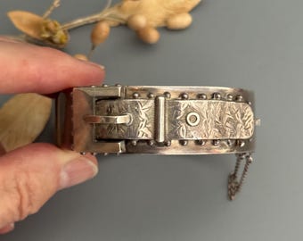 May include: Silver bracelet with a buckle closure and a delicate chain detail. The bracelet is engraved with a floral pattern.