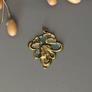May include: A gold-toned pendant with a woman's profile and teal and white enamel accents. The pendant has an ornate, Art Nouveau design with a loop for a chain. The background is a solid gray.