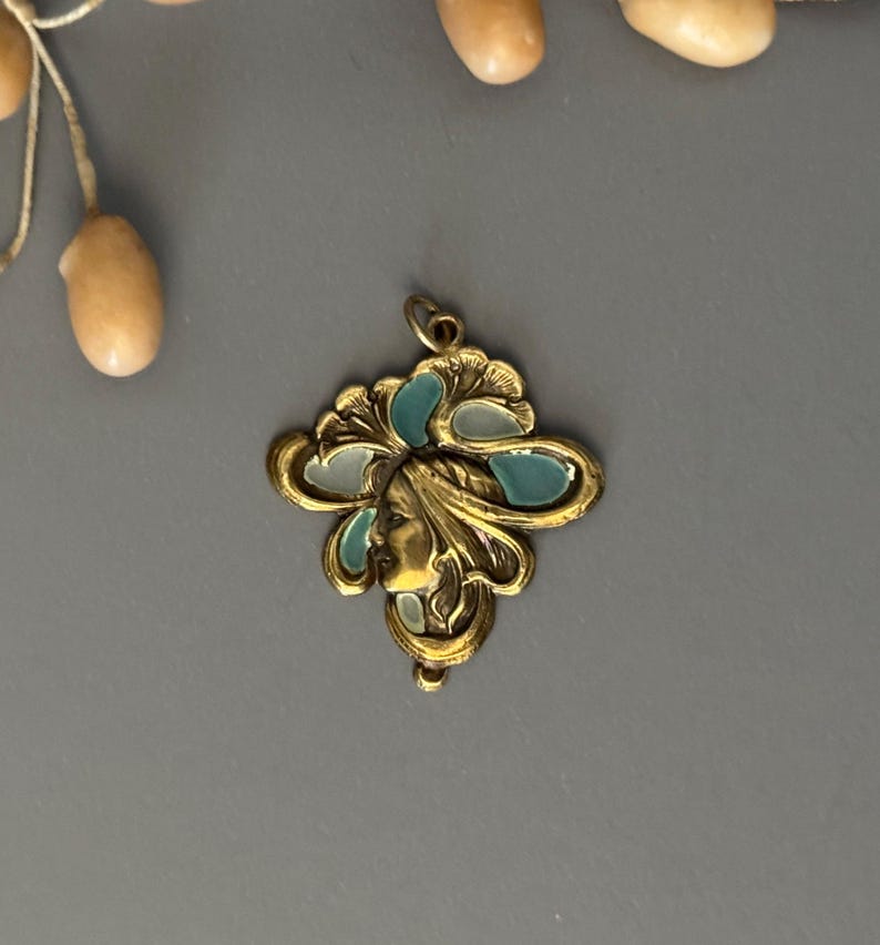 May include: A gold-toned pendant with a woman's profile and teal and white enamel accents. The pendant has an ornate, Art Nouveau design with a loop for a chain. The background is a solid gray.